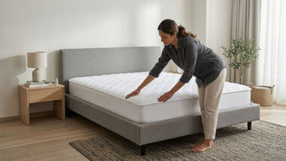 Woman smoothing a waterproof mattress protector on a grey bed frame in a minimalist bedroom